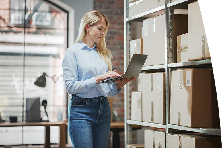 Female Inventory Manager Checks Stock, Writing in Clipboard App on Laptop Computer. Blond Woman Working in a Warehouse Storeroom with Rows of Shelves Full of Parcels, Packages Ready for Shipment.